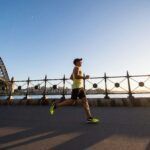 A man is running on the road next to a river. A bridge is visible behind him.